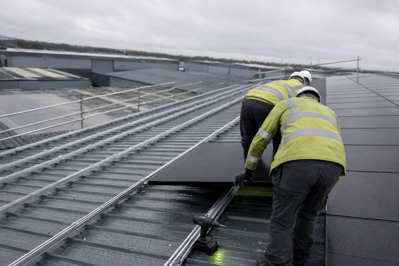 Two installers in hi-vis lowering a black solar module onto aluminium mounting rails on the wet profiled metal roof of a UK industrial unit