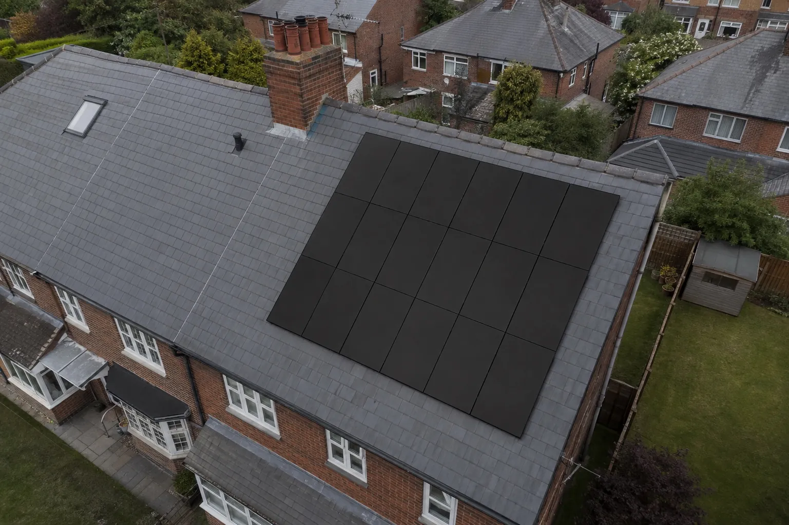 An aerial view of a rectangular array of black solar panels covering one slope of a British slate roof, with the garden and neighbouring houses below