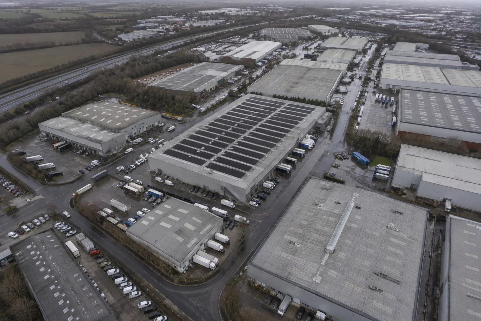 An aerial view over a British industrial estate on an overcast day, one large flat roof covered in rows of matte black solar panels among neighbouring bare roofs