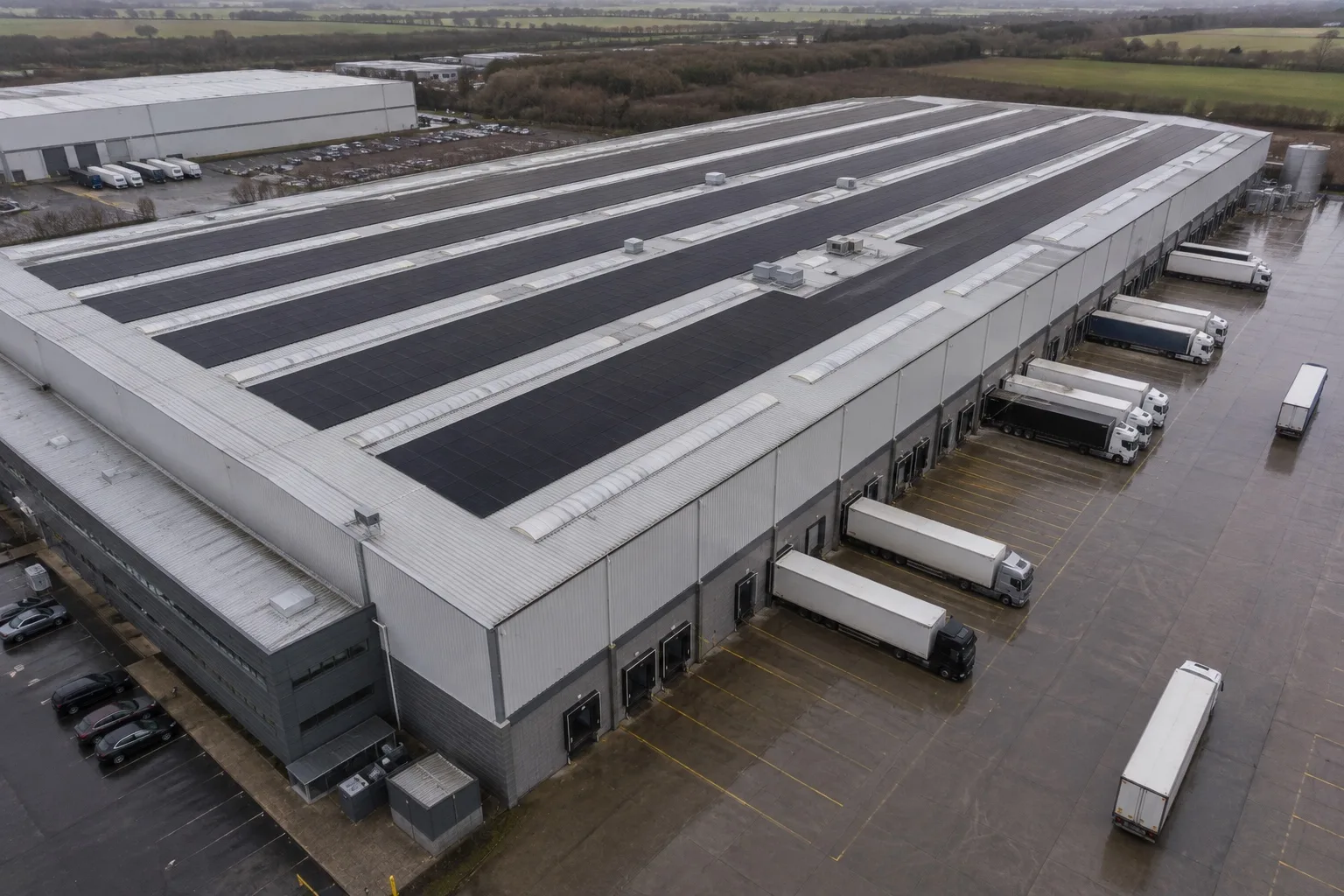 An aerial view of a British logistics warehouse roof carrying long rows of matte black solar panels, with articulated trailers at the loading docks below