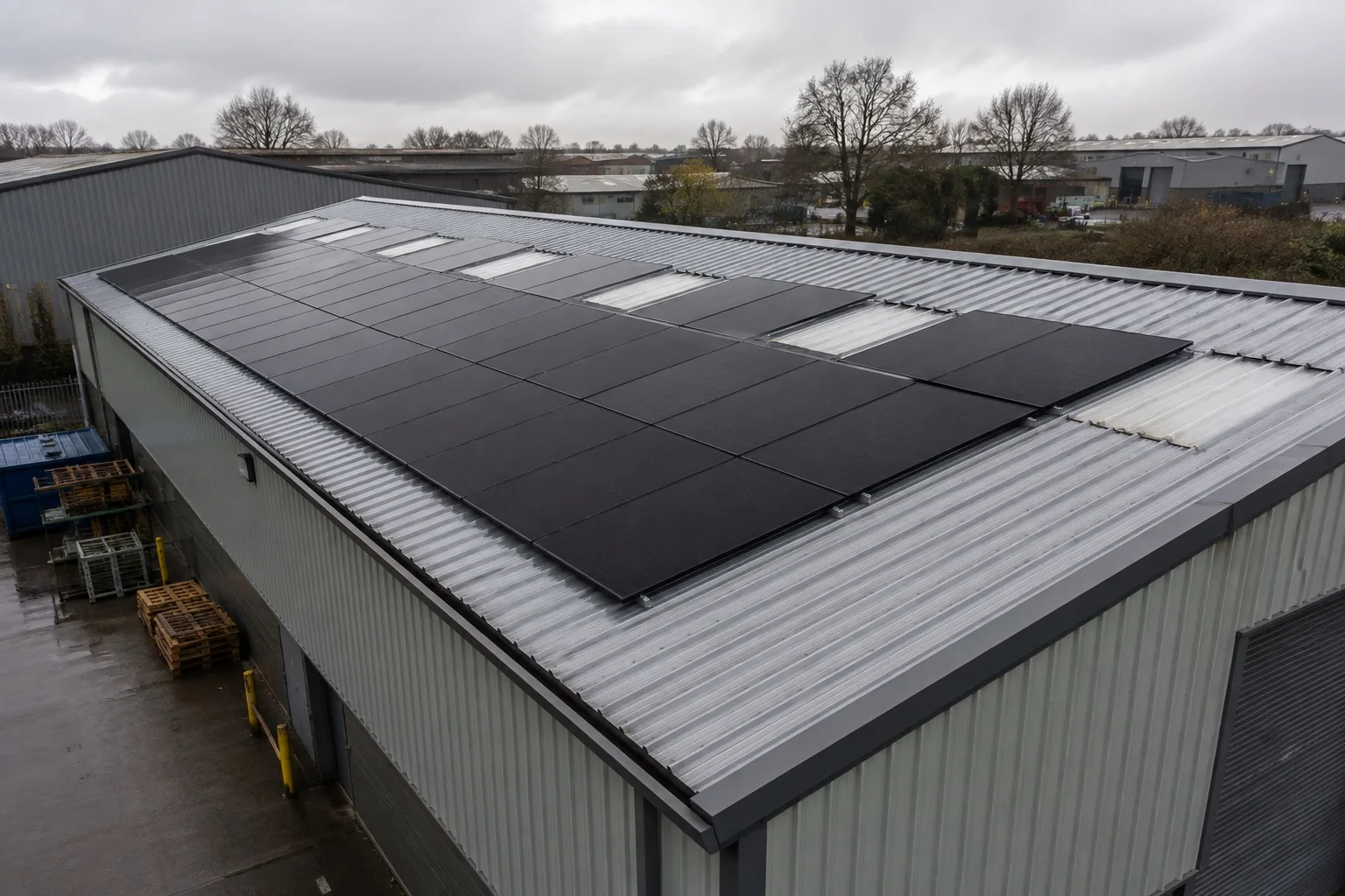 The profiled metal roof of a British industrial unit after rain, carrying matte black solar panels mounted flush to the sheeting