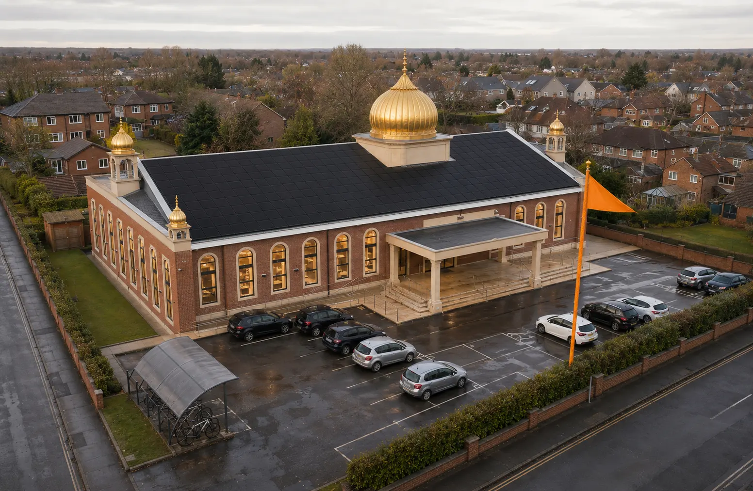 A British gurdwara seen from above in low winter light, black solar panels filling its roof beneath a gilded gold dome, with a saffron nishan sahib on the forecourt