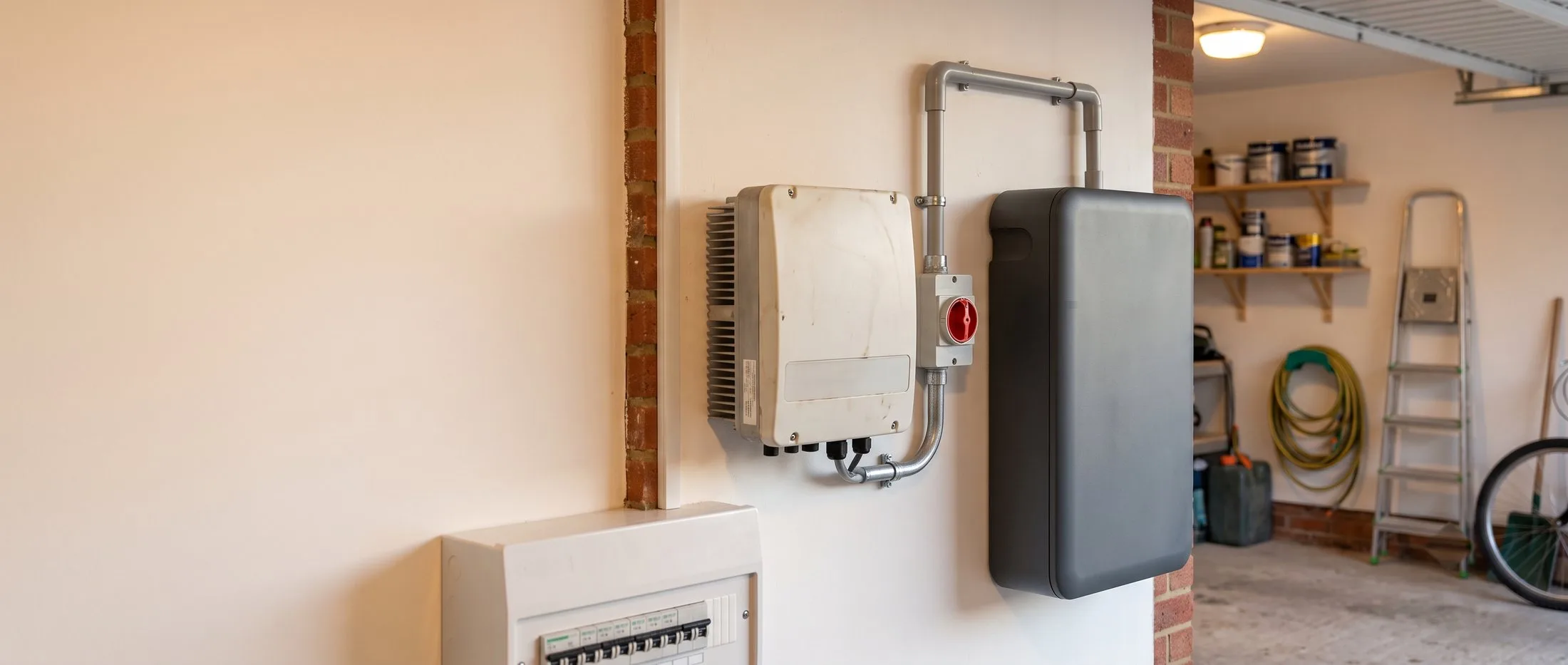 Adding to a system you already own: battery storage beside the existing inverter A newly installed home battery mounted beside an existing solar inverter in a UK garage, joined by conduit through a red isolator