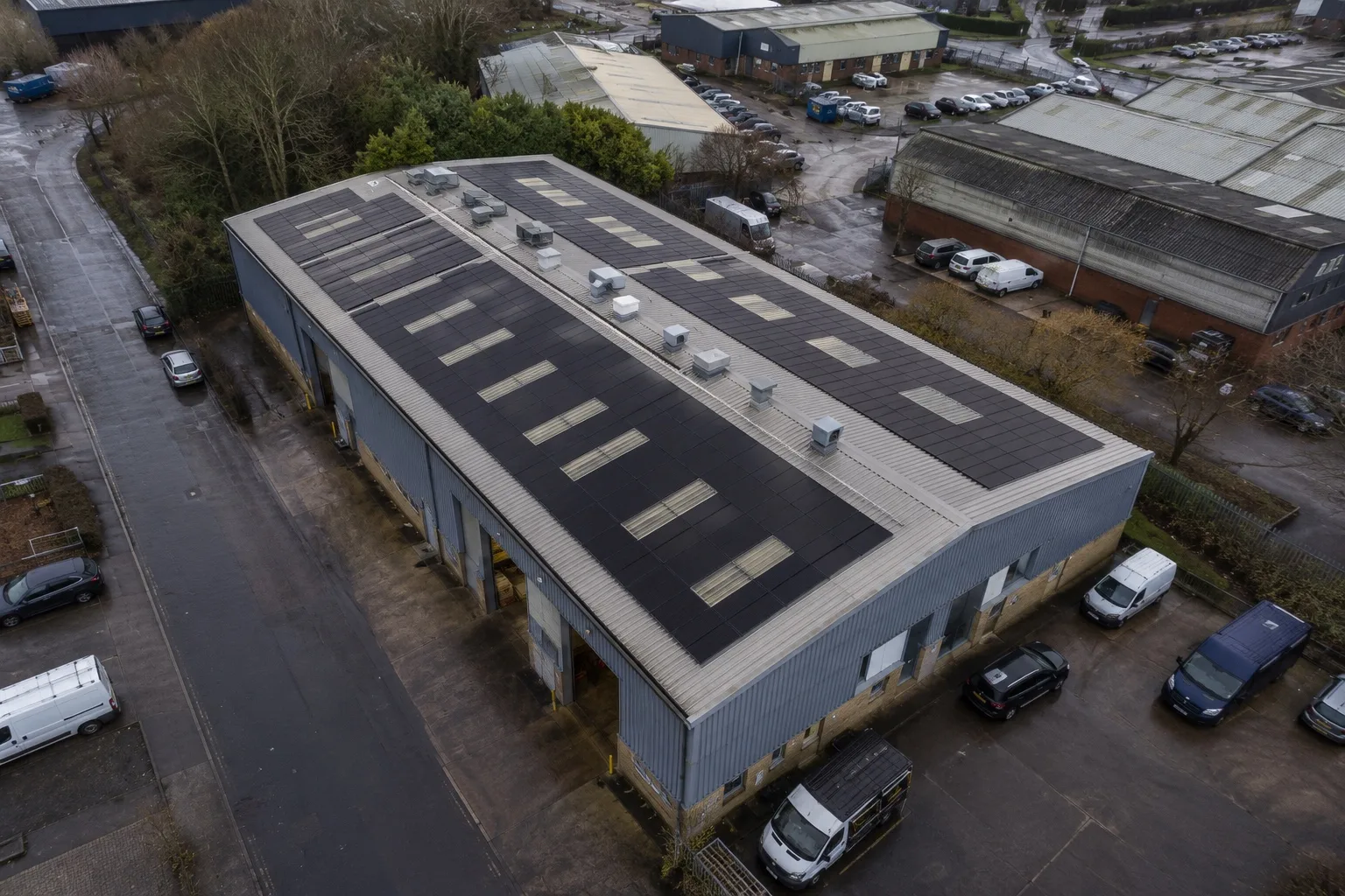 An aerial view of a British trade unit whose roof carries rows of matte black solar panels shaped around the rooflights, with vans parked in the yard